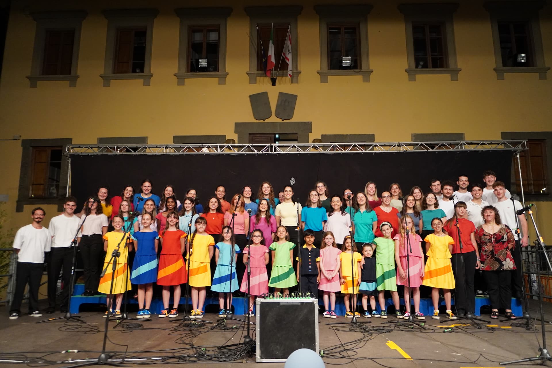 Coro Arcobaleno sul palco, bambini e ragazzi con abiti colorati disposti in file, sfondo scuro e struttura metallica superiore