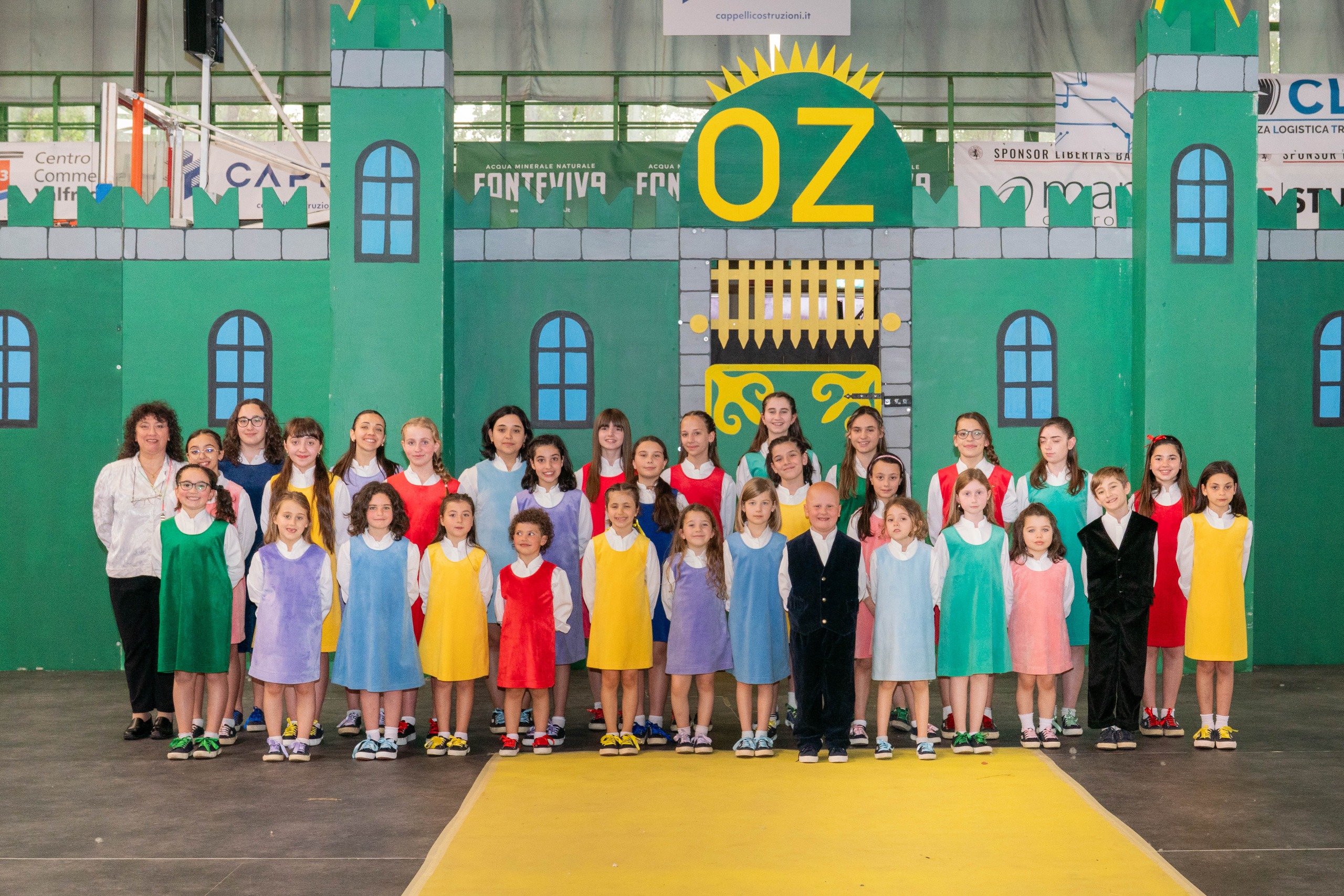 Coro Arcobaleno sul palco, bambini e ragazzi con abiti colorati disposti in file, sfondo scuro e struttura metallica superiore